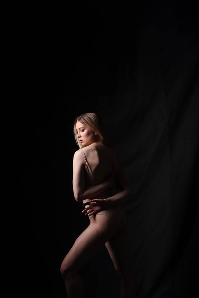 Ballerina in a beige bodysuit. Dark background. Sculpted beautiful female body. Pose of a gymnast. - Foto, imagen