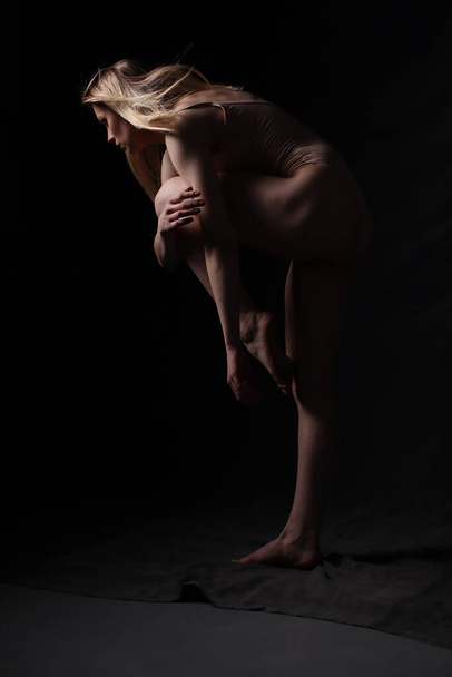 Bailarina en un body beige. Fondo oscuro. Esculpido hermoso cuerpo femenino. Pose de una gimnasta. - Foto, imagen