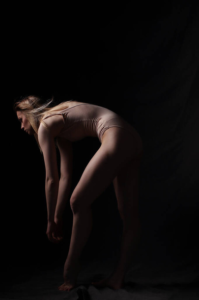 Ballerina in a beige bodysuit. Dark background. Sculpted beautiful female body. Pose of a gymnast. - Фото, изображение