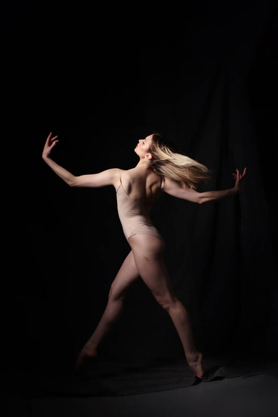 Ballerina in a beige bodysuit. Dark background. Sculpted beautiful female body. Pose of a gymnast. - 写真・画像