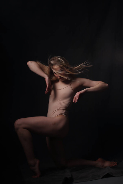 Ballerina in a beige bodysuit. Dark background. Sculpted beautiful female body. Pose of a gymnast. - Fotó, kép