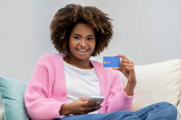 Pozytywny wesoły millenium afrykańskiej pani w stroju casual siedzi na kanapie w domu, trzymając niebieską plastikową kartę bankową i telefon komórkowy, zakupy lub bankowość online, przestrzeń do kopiowania - Zdjęcie, obraz