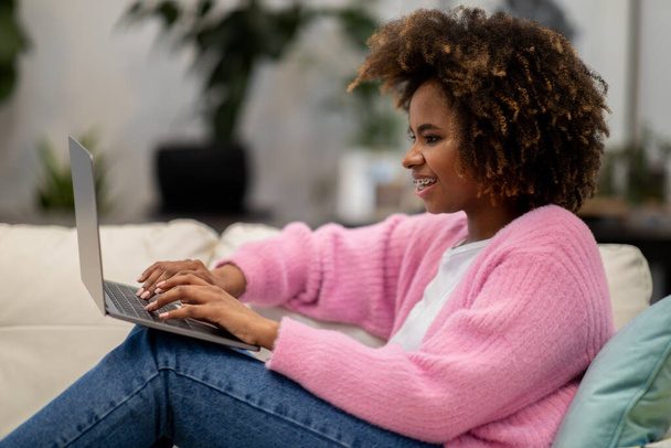 Positive pretty millennial african american lady with bushy hair wearing casual comfy outfit sitting on couch at home, typing on laptop keyboard and smiling, chatting with guys on dating website - Фото, зображення