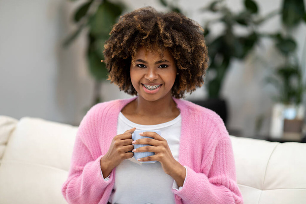 Lachende mooie krullende millennial Afrikaanse Amerikaanse vrouw in comfortabele huiskleding ontspannen op de bank, het houden van koffie mok, genieten van kalmerende kruidenthee, interieur, kopieerruimte - Foto, afbeelding