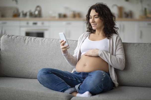 Усміхнена вагітна жінка використовує додаток відстеження вагітності на смартфоні, розслабляючись на дивані вдома, красива майбутня мати читає блог материнства онлайн на мобільному телефоні під час відпочинку на дивані
 - Фото, зображення