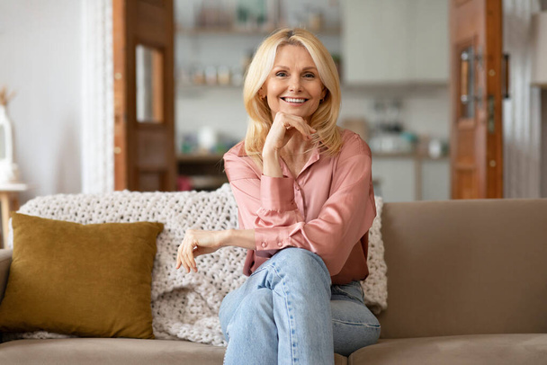 Happy Blonde Mature Woman Enjoying Her Time At Home, Posing Sitting On A Cozy Couch And Smiling To Camera Resting Chin On Hand. Portrait Of Confident Lady. Domestic Comfort Concept - Foto, Imagem
