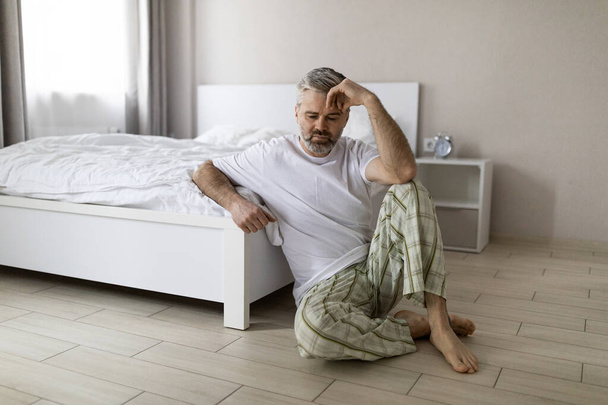 Upset pensive middle aged grey-haired handsome man wearing pajamas sitting on floor by bed at home, touching head, suffering from hangover or anxiety, copy space. Permacrisis, midlife crisis - Foto, Bild