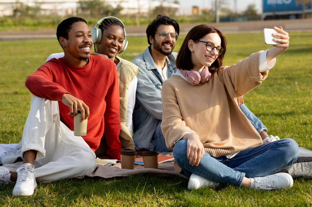 Gelukkige internationale millennial studenten studeren, drinken koffie, ontspannen, zitten op gras, selfie samen outdoor campus. Foto voor blog en sociale netwerken, pauze en onderwijs, communicatie - Foto, afbeelding