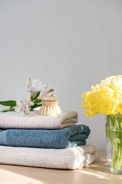 Vertical shot with stack of dry clean and soft towels, small wooden brush and flowers on the beige shelf in room. Bouquet with tulips in glass vase near bathroom textile - Photo, Image