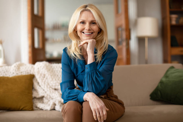 Selbstbewusste Blonde Reife Geschäftsfrau posiert mit anrührendem Kinn auf dem Sofa im Haus und drückt positive Emotionen aus. Professionelle Psychologin im mittleren Alter lächelt in die Kamera im Büro - Foto, Bild