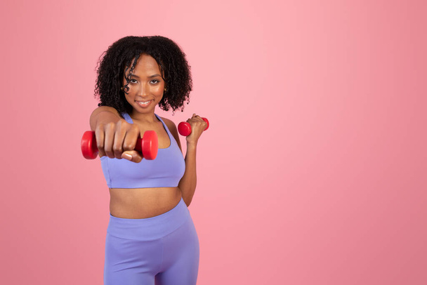 Χαρούμενα χιλιετή αφρικάνικη αμερικανική κυρία στο αθλητικό ντύσιμο απολαύσετε ασκήσεις με τα χέρια αλτήρες απομονώνονται σε ροζ φόντο, στούντιο. Αθλητική διαφήμιση και προσφορά, pilates, fitness και φροντίδα του σώματος, κίνητρα - Φωτογραφία, εικόνα