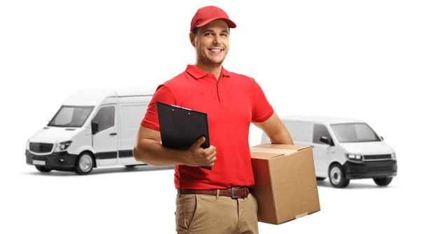 Delivery worker in a uniform holding a clipboard and a box in front of transport vans isolated on white background - Fotó, kép