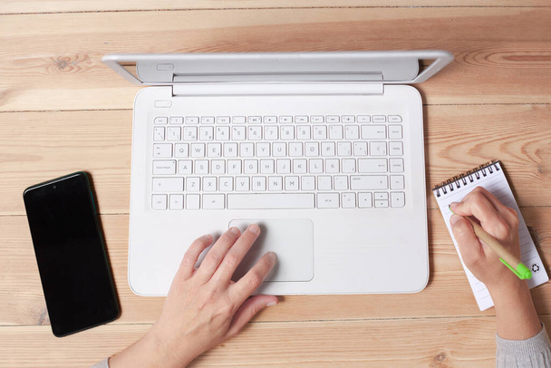 Une femme est vue travaillant à la maison, contrôlant le trackpad de son ordinateur portable d'une main tout en écrivant dans un bloc-notes avec l'autre. - Photo, image