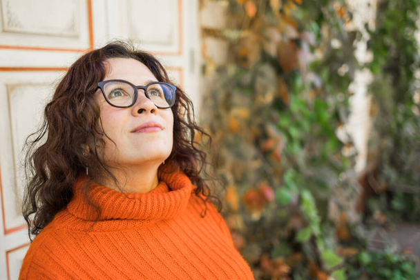 Autumn portrait of an attractive young woman in stylish glasses in a knitted fashionable orange sweater on background leaves. Girl walks in city - Valokuva, kuva