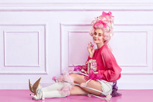 Elegante dama. Retrato de niña, princesa con ropa rosa de lujo sosteniendo caramelos sobre fondo interior de lujo. Concepto de comparación de épocas, modernidad y renacimiento, belleza, historia - Foto, imagen