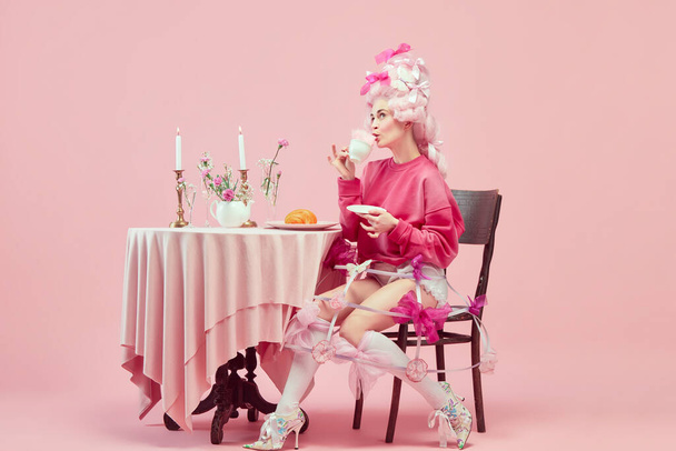 Royalty breakfast. Portrait of elegant princess, queen wearing pink clothes, and wig drinking coffee over pink background. Concept of comparison of eras, modernity and renaissance, beauty, history - Photo, Image
