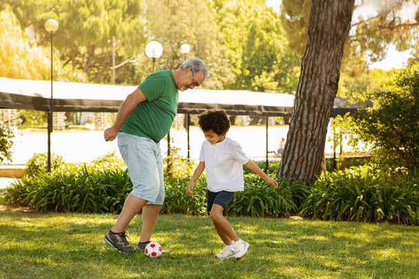 Cheerful little mixed race boy and old caucasian grandfather enjoy free time, have fun, playing soccer ball outdoor. Football in park together, family relationships, weekends and hobbies - Fotó, kép