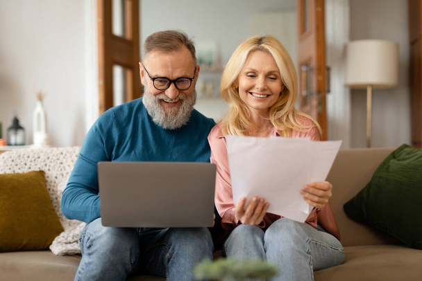 幸せな成熟したカップルは紙仕事をし、ノートパソコンを使用して、オンライン財務を管理し、自宅で紙の請求書と通信を読んで、眼鏡をかけています。家庭文書概念 - 写真・画像