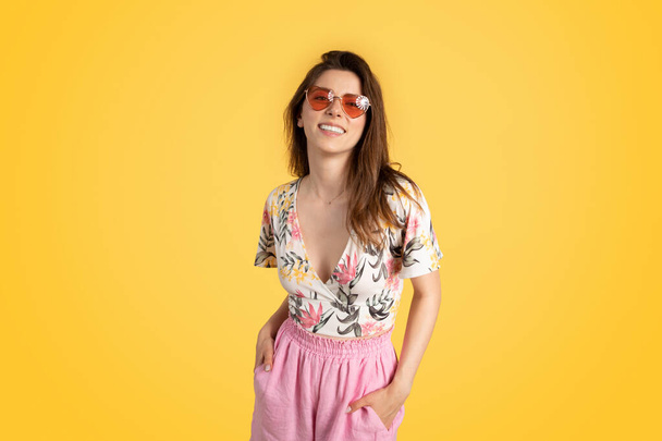 Geniet van de zomer. Mooie jongedame in trendy zomerkleding en kleurrijke zonnebril, lachend naar de camera, poserend op een gele studio achtergrond - Foto, afbeelding