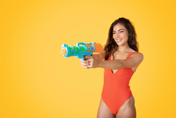 Glad millennial asian lady in swimsuit with water gun shooting on empty space isolated on orange studio background. Pool and sea fun, enjoy summer, rest and relax, game killer - Foto, Imagem