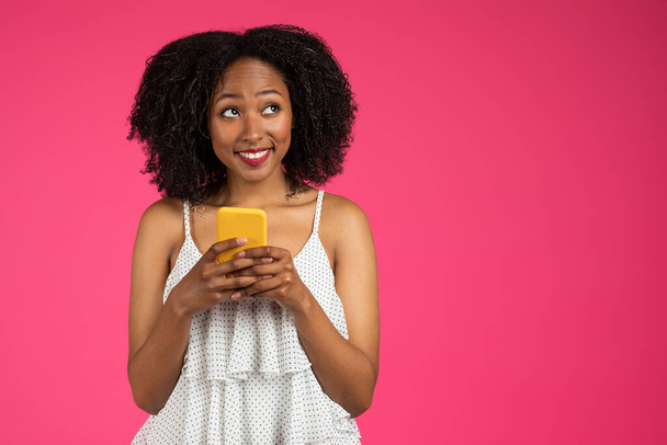 Uśmiechnięta młoda afrykańska kręcona kobieta rozmawiająca na smartfonie, patrząca na puste miejsce odizolowane na różowym tle studia. Romantyczna wiadomość, social media, blog i gadżet, reklama, oferta - Zdjęcie, obraz