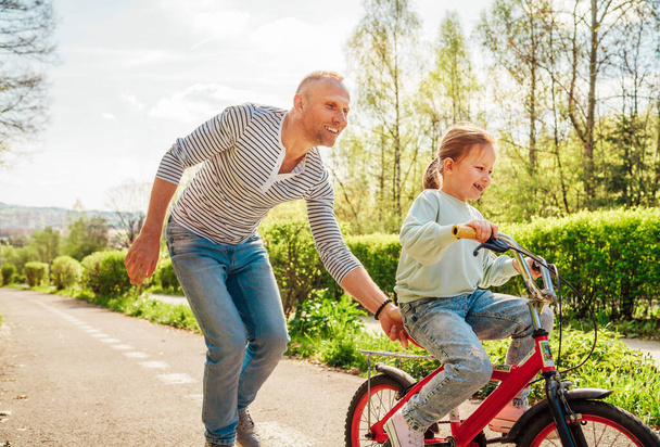Smiling father teaching his daughter to ride a bicycle. They enjoy togetherness in the summer city park. Happy childhood and outdoor activities concept image. - Photo, image