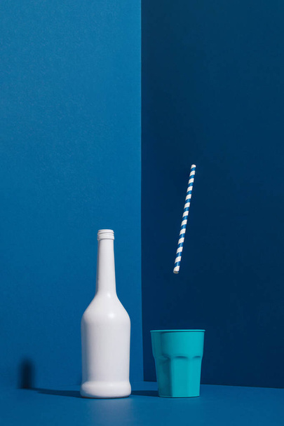 青い壁の隅にプラスチック製のカップに落ちる剥がれた藁と白のドリンクボトル。ミニマルな夏のコンセプト. - 写真・画像
