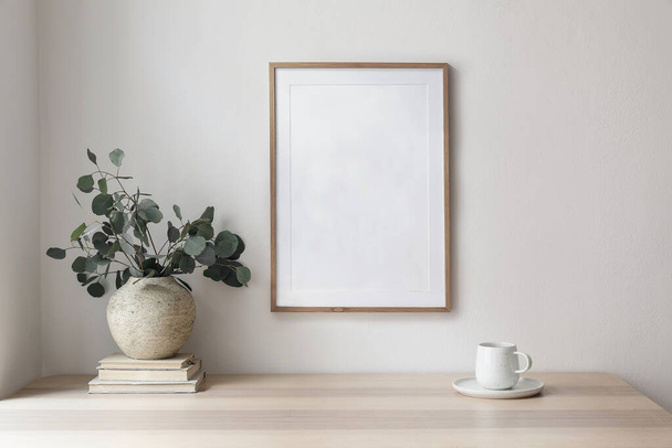 Empty wooden picture frame, poster mockup hanging on beige wall background. Vase with green eucalyptus tree branches on table. Cup of coffee, books. Working space, home office, modern art display. - Foto, Imagen