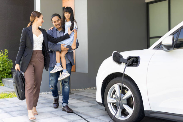 Progressive young parents and daughter with electric vehicle and home charging station. Green and clean energy from electric vehicles for healthy environment. Eco power from renewable source at home. - Foto, Bild