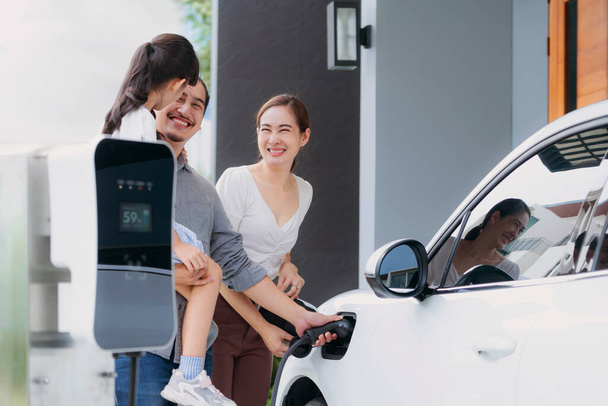 Progressive young parents and daughter with electric vehicle and home charging station. Green and clean energy from electric vehicles for healthy environment. Eco power from renewable source at home. - Valokuva, kuva