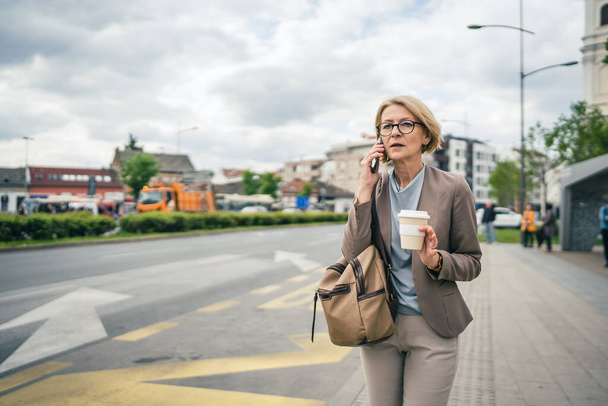 One woman caucasian mature blonde female with eyeglasses stand at bus city stop in day use smartphone mobile phone making a call talk while wait receive bad news - Photo, Image