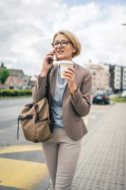 One woman caucasian mature blonde female with eyeglasses stand at bus city stop in day use smartphone mobile phone making a call talk while wait for the drive real person copy space late for work - Photo, Image