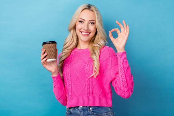 Photo of good mood pretty girl with wavy hairdo dressed pink sweater showing okey hold cup of latte isolated on blue color background. - Photo, Image