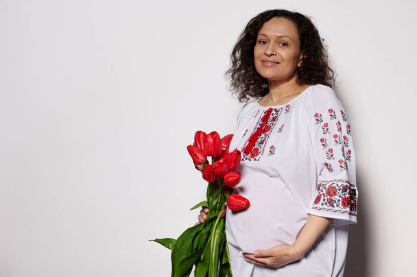 Happy delightful pregnant woman holding a bouquet of red tulips, caressing her belly, smiling, looking at camera on isolated white background, dressed in traditional ukrainian embroidered white dress - Photo, image