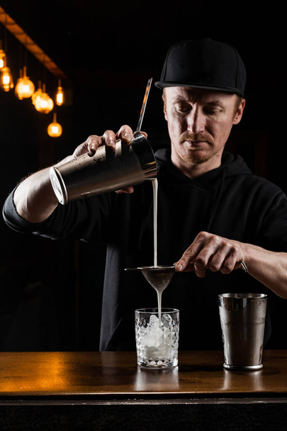 Barman schenkt gemengde Penicilline cocktail in het glas aan de bar. Barman mengt whisky, citroensap en honing-gember siroop om de Penicilline alcoholische cocktail te bereiden - Foto, afbeelding