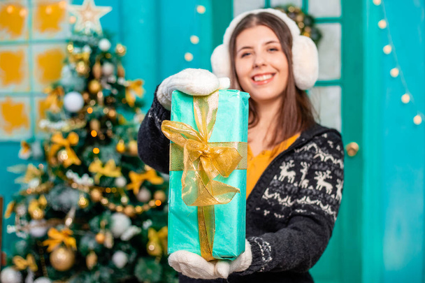 Close-up of a gift in a turquoise wrapper with a golden bow in the hands of a young woman dressed in a sweater and mittens, posing standing against the backdrop of a Christmas tree. - Foto, Bild