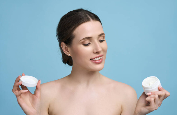 Smiling young Caucasian woman holding cosmetics mockup jar, facial cream template jar. Pretty female with healthy glowing skin and natural makeup, posing bare shoulders on isolated blue background - Фото, зображення