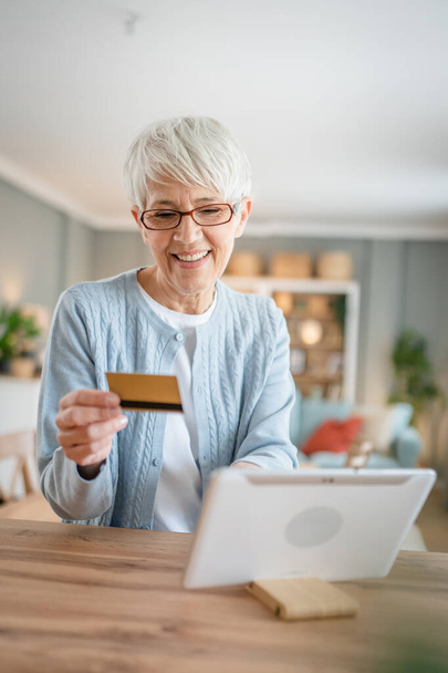 Jedna dojrzała starsza kobieta babcia siedzieć w domu używać karty kredytowej lub debetowej do zakupów online przeglądać sklepy internetowe kupowanie rzeczy prawdziwych ludzi kopiować przestrzeń - Zdjęcie, obraz