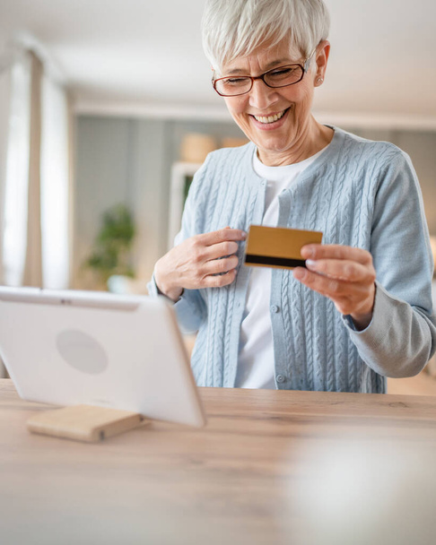 ある成熟した年配の女性の祖母はオンラインショッピングのための家の使用のクレジットカードかデビットカードに坐る実際の人々がスペースをコピーするものを買うインターネット店 - 写真・画像