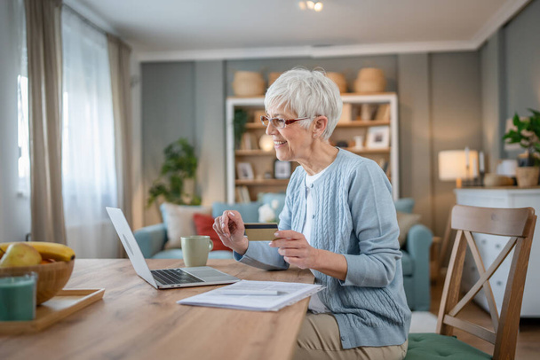 Yetişkin bir yaşlı kadın büyükanne evde oturup online alışveriş siteleri için kredi kartı ya da banka kartı kullanıyor. İndirimli indirimli ürünler alıyor. Gerçek insanlar bilgisayar kopyalıyor. - Fotoğraf, Görsel
