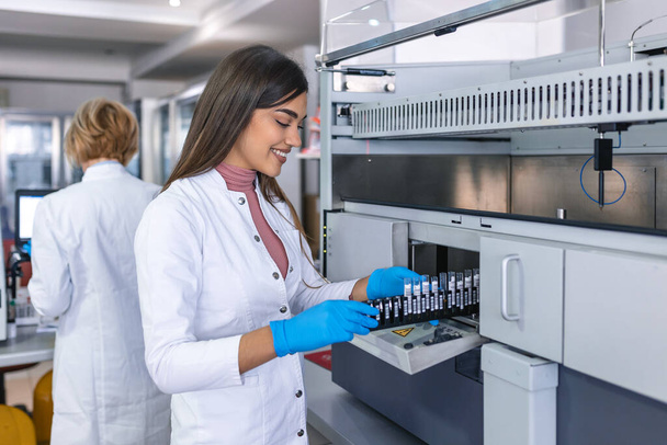 Portrait of a young female laboratory assistant making analysis with test tubes and analyzer machines sitting at the modern laboratory - Foto, afbeelding