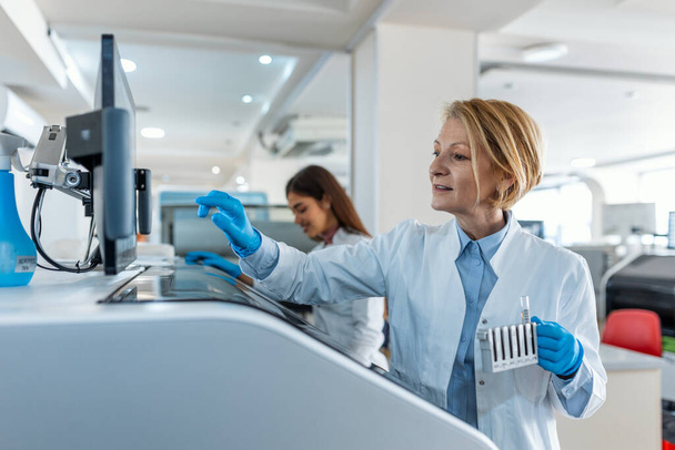 Portrait of a young female laboratory assistant making analysis with test tubes and analyzer machines sitting at the modern laboratory - Фото, изображение
