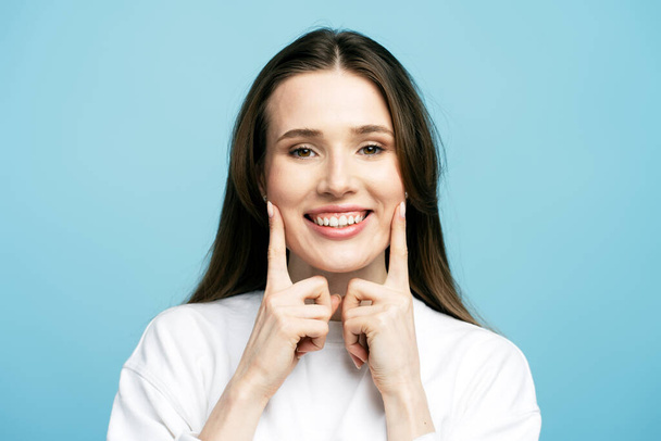 Close-up portret van gelukkige mooie jonge vrouw met tanden glimlach wijzend met vingers op zijn mond op zoek naar camera geïsoleerd op blauwe achtergrond. Gezondheidszorg, kliniek, tandheelkundige behandeling concept - Foto, afbeelding