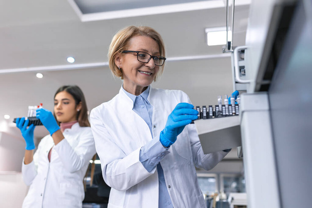 V laboratoři ženské vědec kontroluje lékařské zařízení analýzy zkumavky s krevními vzorky. Tým výzkumných pracovníků ve farmaceutické laboratoři. - Fotografie, Obrázek