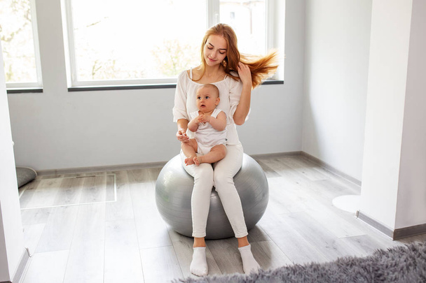 Ευτυχισμένη μητέρα κρατώντας το κοριτσάκι, ενώ γερός σε μπάλα γυμναστικής στο άνετο σπίτι. Ασκήσεις μπάλα σταθερότητας για τη νευροανάπτυξη του μωρού. - Φωτογραφία, εικόνα