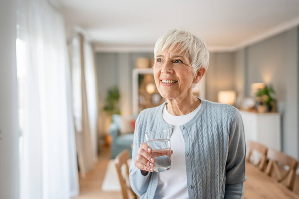 Közelkép portré egy idős nő rövid haj boldog mosoly pozitív érzelem másolat tér áll otthon beltéri szürke fehér haj hold pohár víz - Fotó, kép