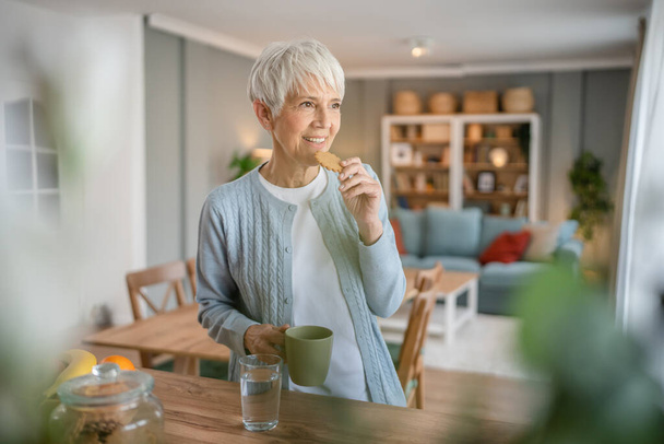 Close up portret jednej starszej kobiety z krótkimi włosami szczęśliwy uśmiech pozytywne emocje przestrzeń ksero stoi w domu kryty szary białe włosy jedząc śniadanie lub przekąskę trzymać ciasteczko i filiżankę kawy - Zdjęcie, obraz