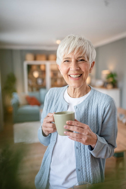 Nahaufnahme Porträt einer älteren Frau mit kurzen Haaren glücklich lächeln positive Emotionen Kopierraum zu Hause stehend grau weiße Haare halten Tasse Kaffee tägliche Morgenroutine - Foto, Bild