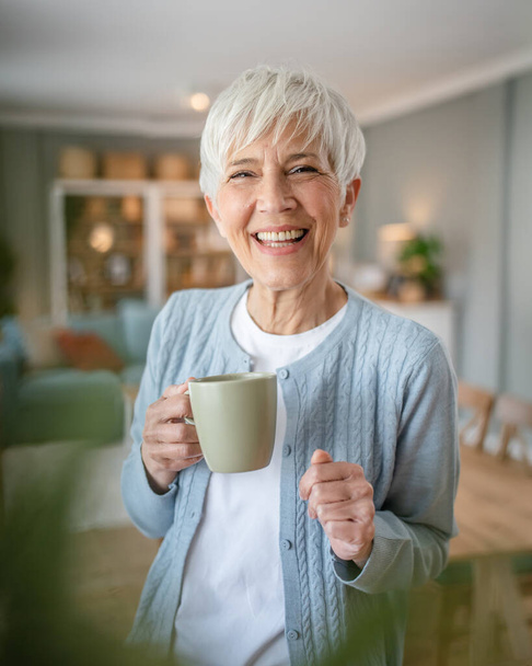 Gros plan portrait d'une femme âgée aux cheveux courts heureux sourire positif émotion copie espace debout à la maison intérieure grise cheveux blancs tenir tasse de café routine quotidienne du matin - Photo, image