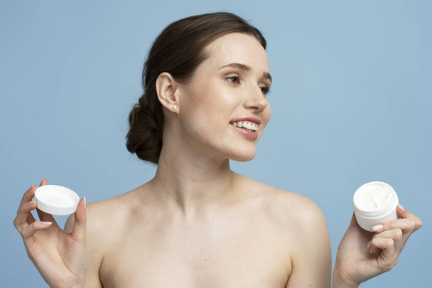 Portrait of beautiful smiling woman holding jar with body cream looking away isolated on blue background. Beauty, skin care, morning routine concept - Φωτογραφία, εικόνα